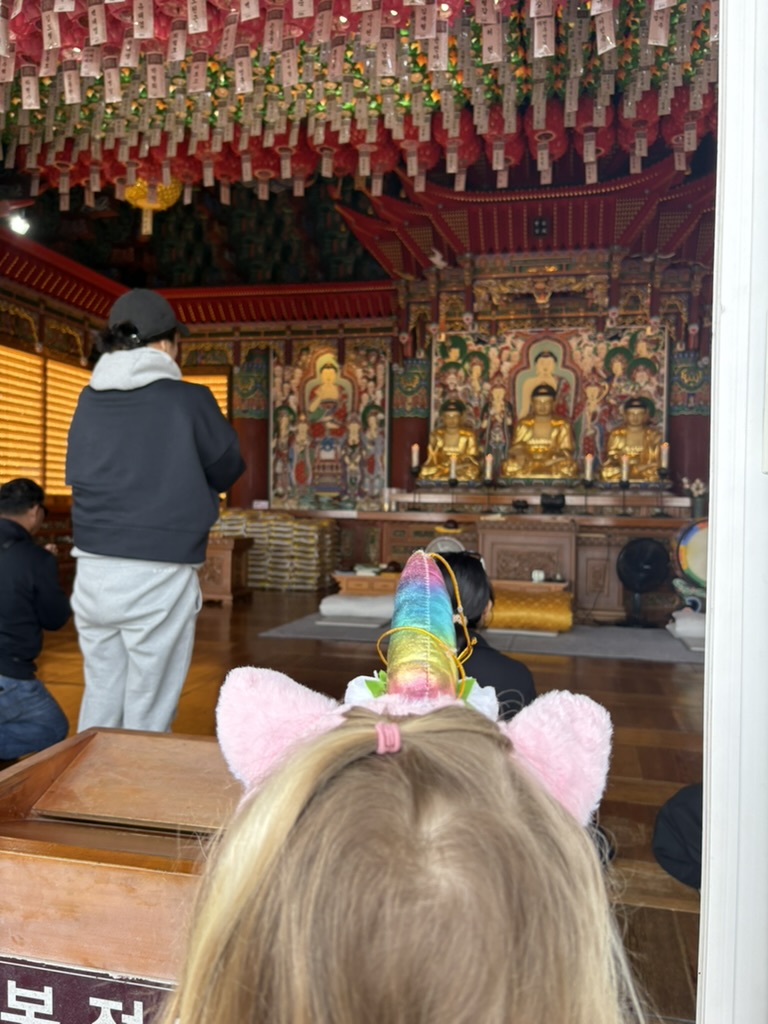 Girl with unicorn headband peering into the ornate temple interior with gilded Buddha statues