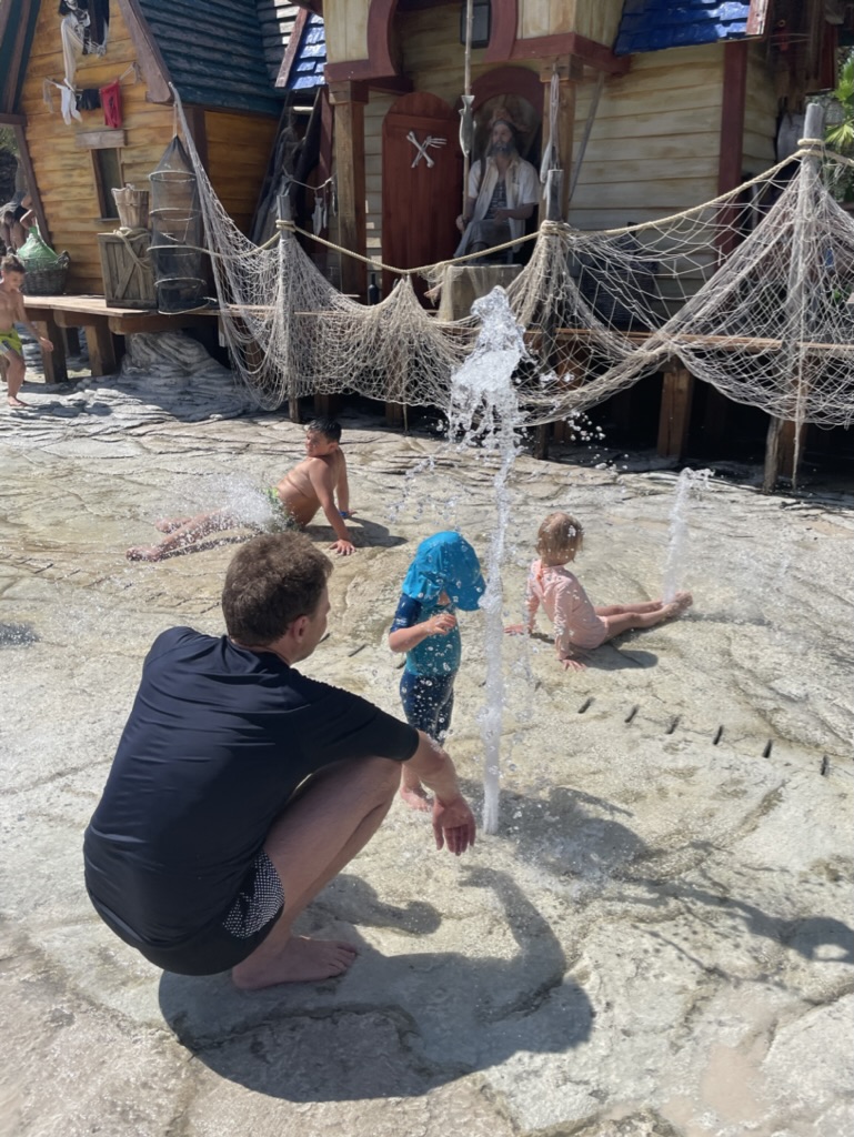 Young children playing in the shallow splash fountains at Caribe Bay's kids' area, with a parent crouching beside them and pirate-village huts behind