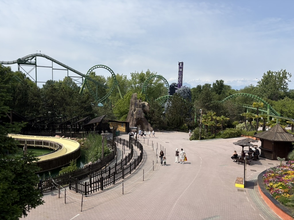 Gardaland park seen from above — green roller coasters, purple drop tower, and the Alps in the distance