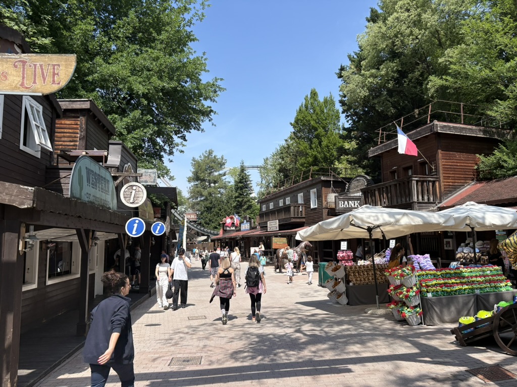 Western-themed area inside Gardaland — wooden buildings, bank sign, market stalls, tall trees