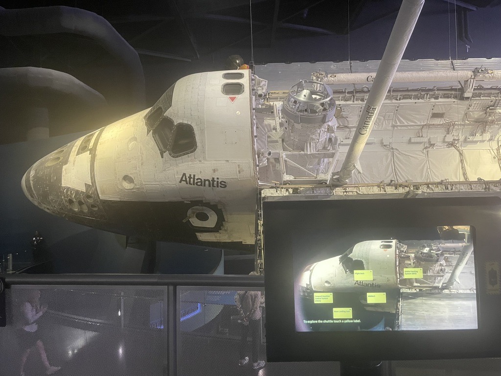 The nose and cockpit of Space Shuttle Atlantis on display at Kennedy Space Center, with the robotic arm extended and an interactive touchscreen in the foreground
