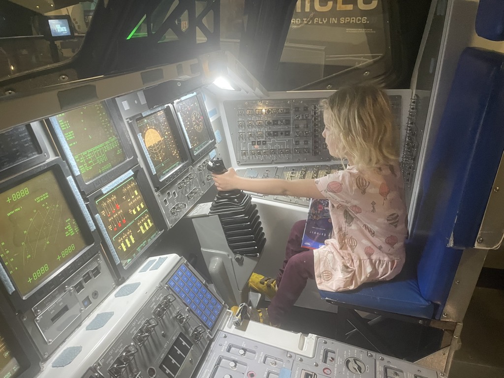A young child sitting in a space shuttle cockpit simulator at Kennedy Space Center, gripping the control stick surrounded by glowing instrument panels