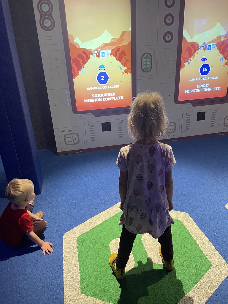 Two young children playing an interactive Mars rover game on a large screen at Kennedy Space Center, standing on a marked floor pad to control the rover