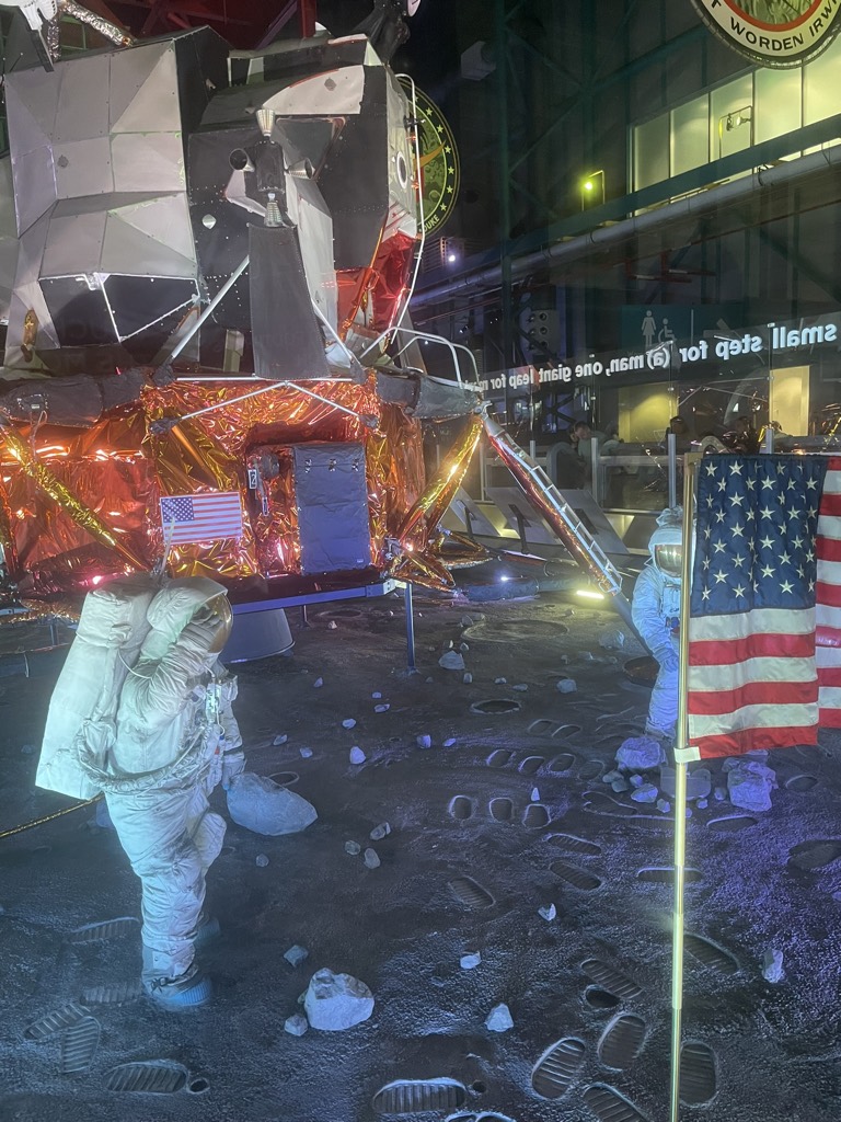 A recreated moon-landing scene at Kennedy Space Center with the lunar module, two astronaut figures in spacesuits, the planted American flag and footprints in the grey lunar surface