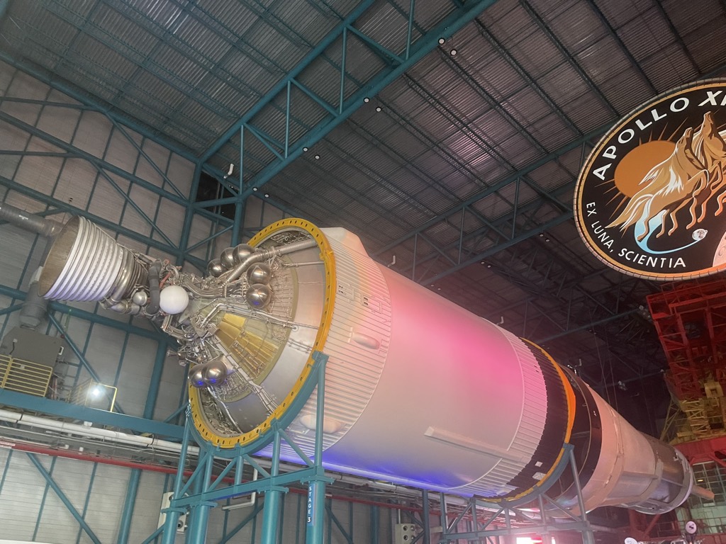 A stage of the Saturn V rocket displayed horizontally inside the Apollo/Saturn V Center at Kennedy Space Center, lit in pink and purple, with an Apollo mission badge on the wall