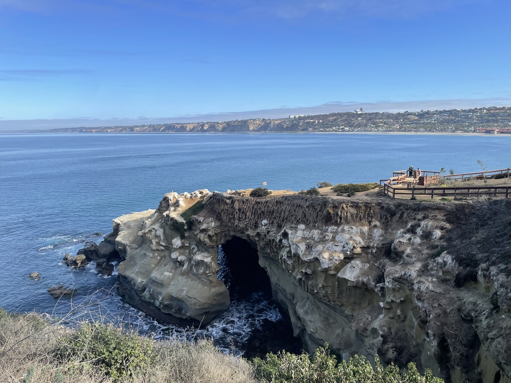 A natural rock arch on the La Jolla cliffs, with the blue Pacific Ocean and coastline stretching behind it and a viewing platform visible above