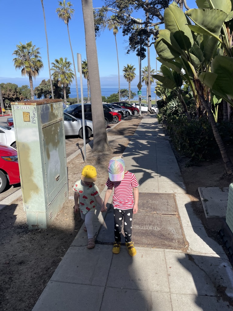 Two young children holding hands on a La Jolla sidewalk, palm trees and the ocean visible ahead of them