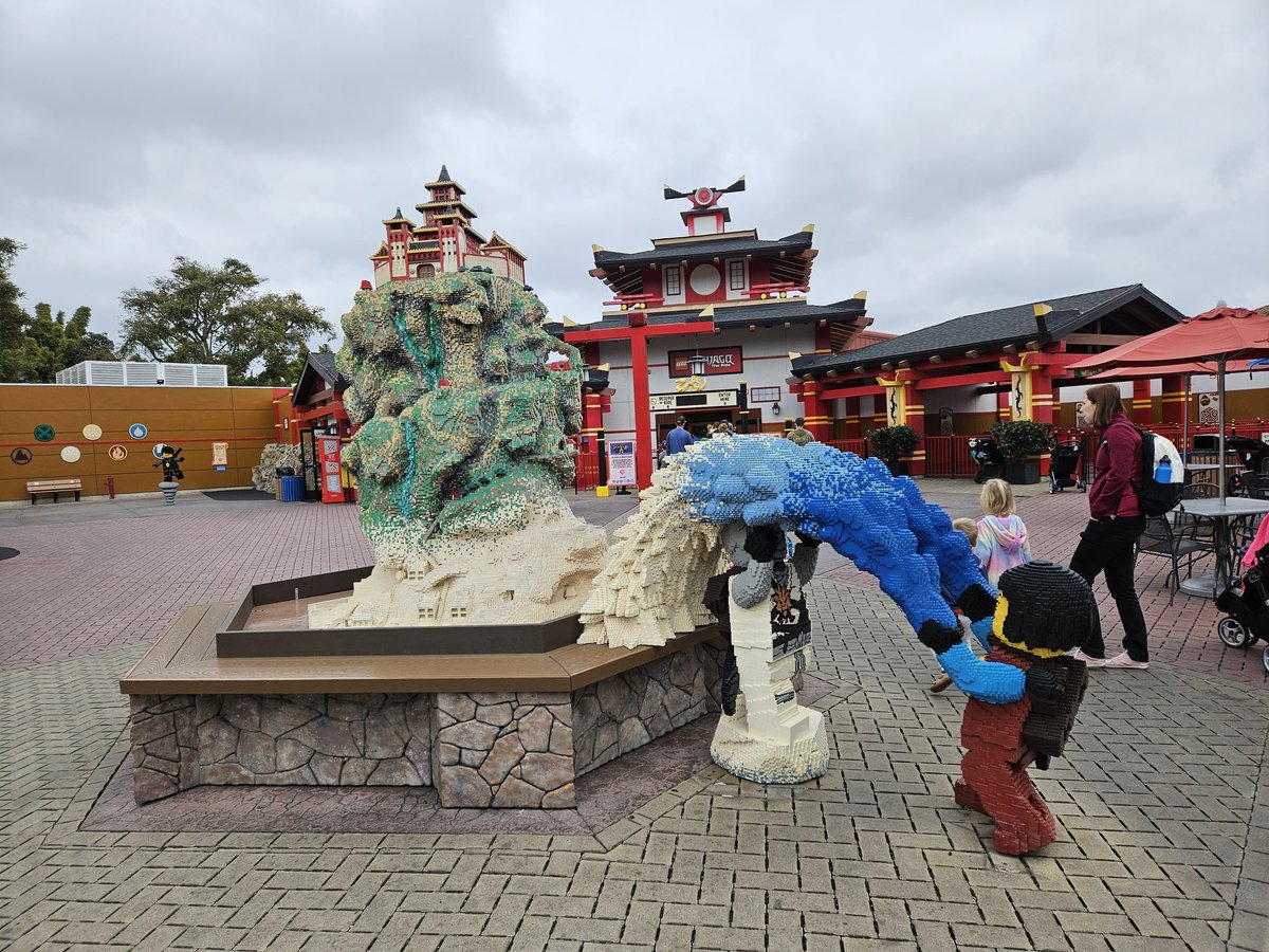 Child leaning over a large LEGO dragon sculpture in the Ninjago area of Legoland California, with the Chinese-style pagoda building behind