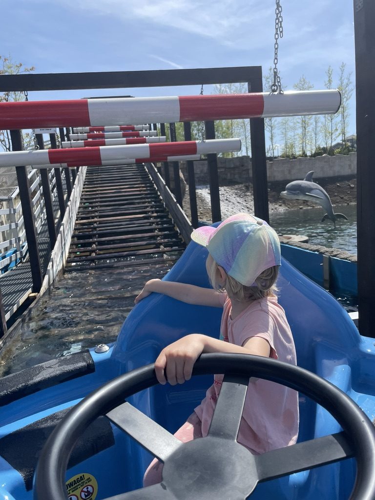 Child in a rainbow cap steering a blue paddle boat on a water ride at Energylandia, dolphin statue in the background