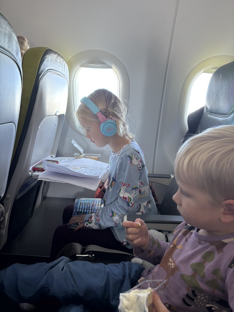 Two young kids on the plane to the Azores, one wearing headphones and coloring, the other eating a snack