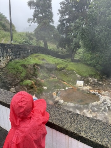 Child in a red raincoat looking at a bubbling volcanic mud pool at Furnas, Azores, in the rain