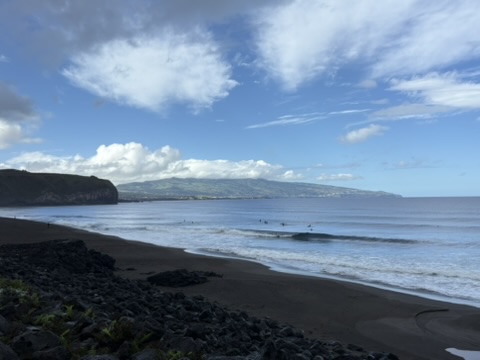 Black sand beach near Ponta Delgada, Azores, with surfers in the water and the island coastline in the distance