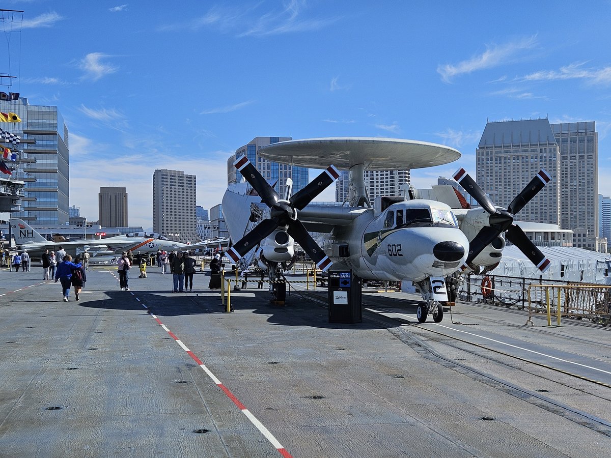 A propeller aircraft on the USS Midway flight deck with the San Diego downtown skyline visible behind it