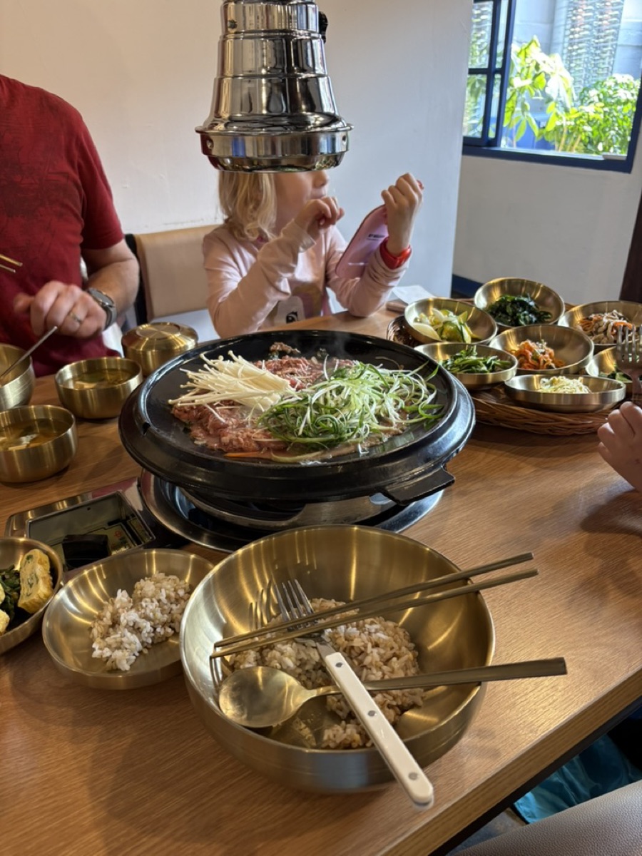 Korean BBQ grill with meat and spring onion, brass bowls of rice and banchan side dishes, and a young child at the table