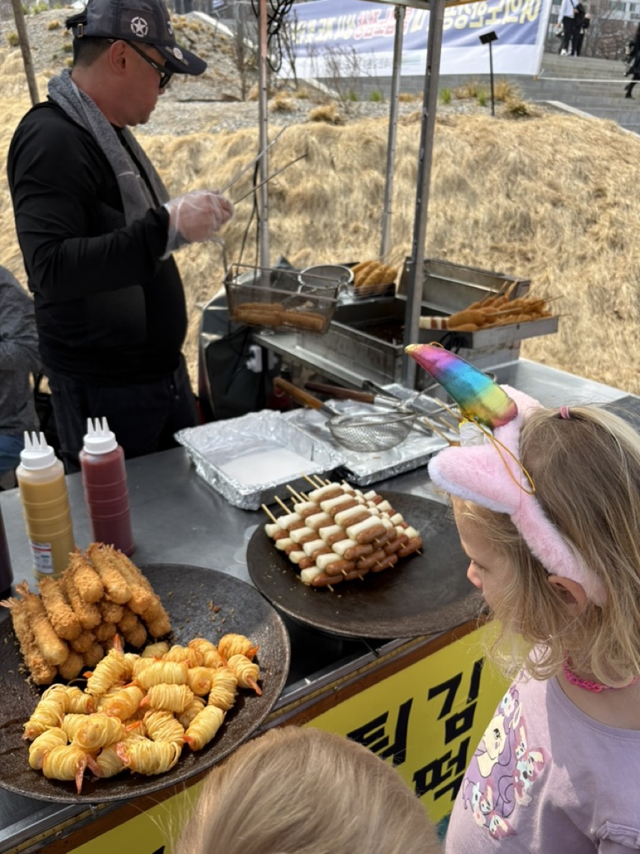 A Korean street food cart with tempura and sausage rice-cake skewers, a child watching