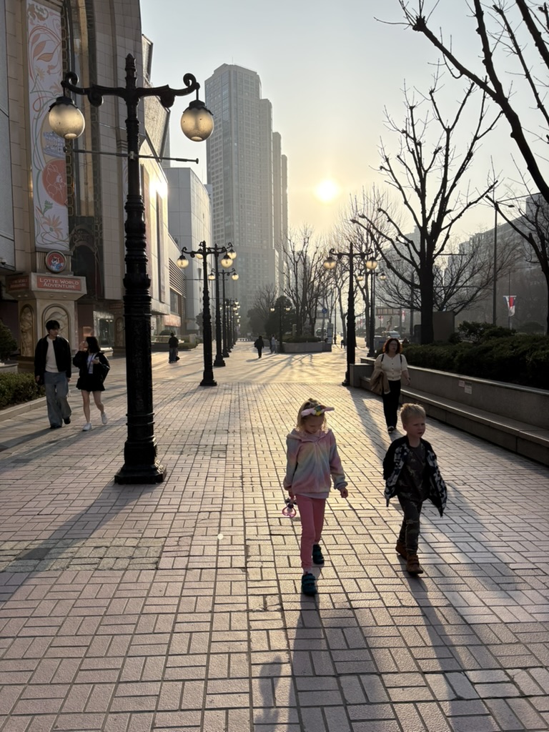 Two kids walking along the wide pedestrian boulevard outside Lotte World Adventure at golden hour, Jamsil