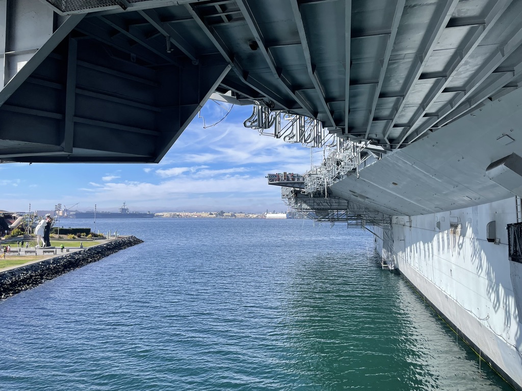 View from beneath the USS Midway flight deck overhang looking out over San Diego Bay, with the Unconditional Surrender statue visible on the shoreline