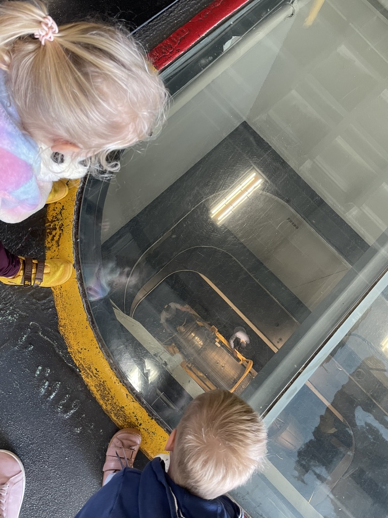 Two children peering through a glass floor panel on the USS Midway flight deck, looking down into the ship below
